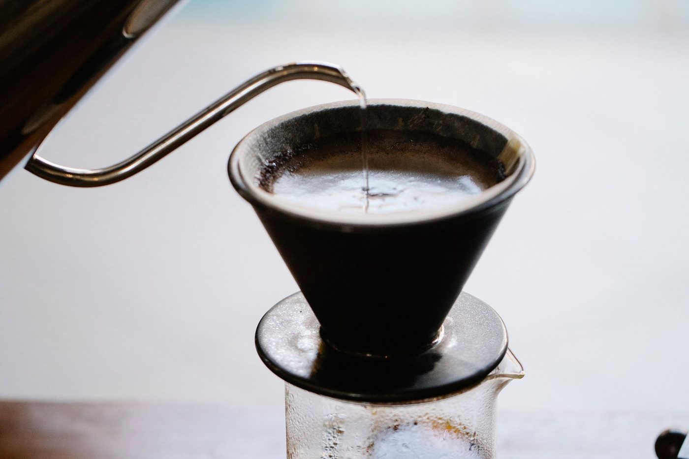 A gooseneck kettle pours a thin stream of water onto a bed of coffee in a dark ceramic dripper set over a glass carafe.