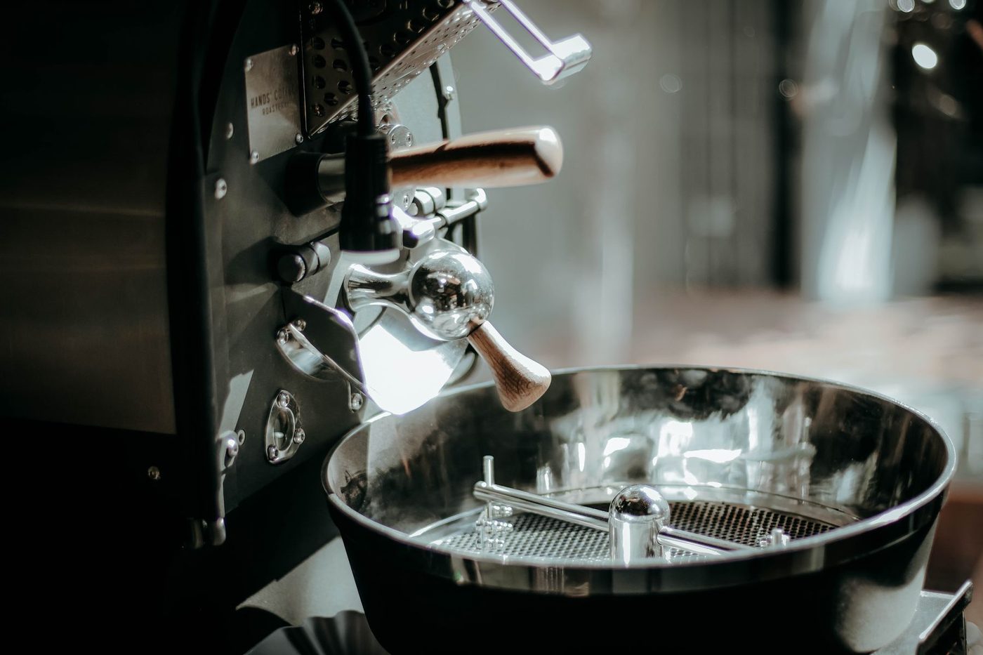 The front of a small drum roaster: the bean chute with its wooden handle, the trier, and the empty cooling tray below.